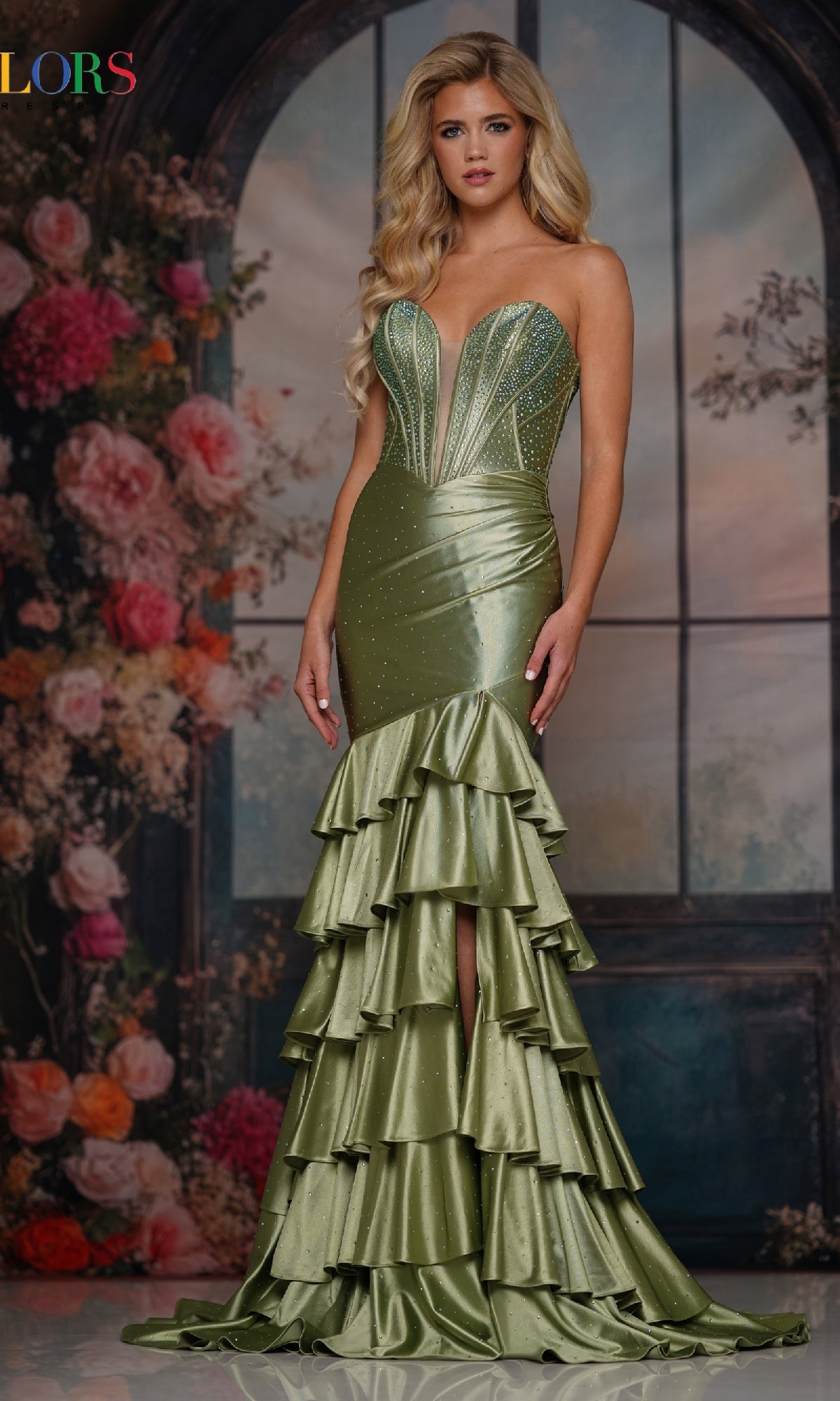 A woman stands before a floral backdrop wearing the Colors Dress 3925, a long prom dress featuring rhinestone embellishments, tiered ruffles, sparkly accents, and a fitted strapless design.